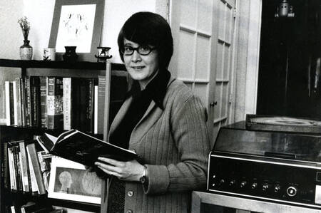 Former Mundelein administrator and teacher stands in front of bookshelf, holding an open book. 